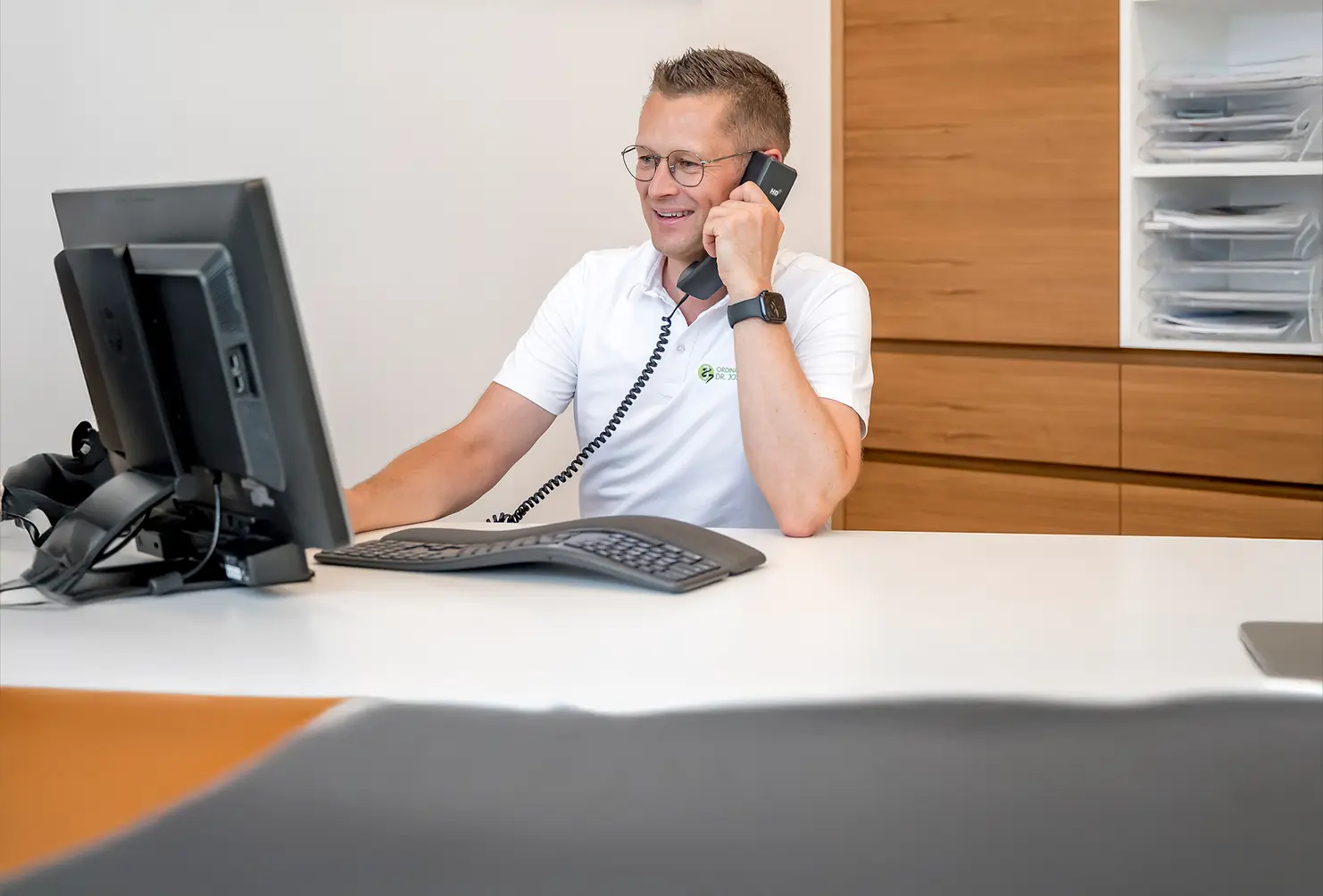 Ein Mann in einem weißen Hemd sitzt an einem Schreibtisch, telefoniert und sieht in Richtung PC-Monitor.