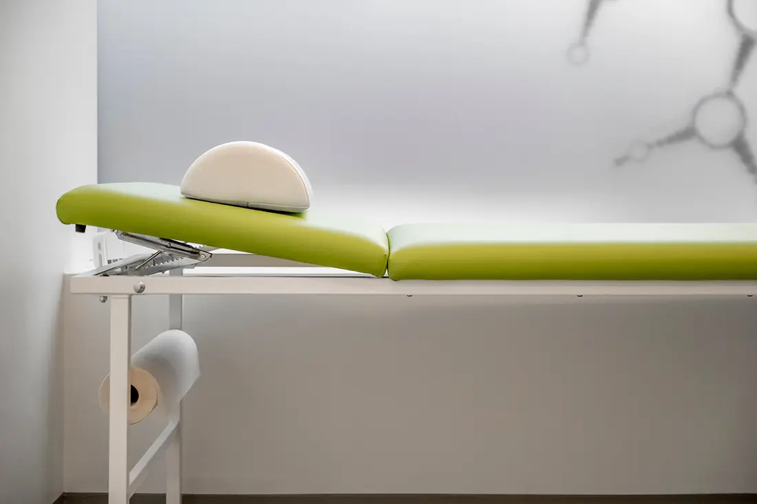 Grün gepolsterter Untersuchungstisch mit weißem Kissen in einem sauberen, modernen Behandlungszimmer.