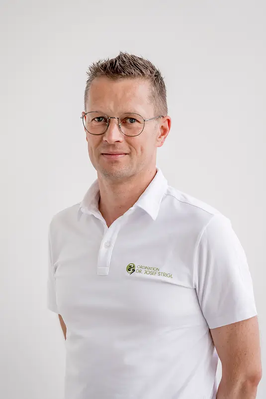 Ein Mann mit Brille und weißem Poloshirt steht vor einem schlichten weißen Hintergrund und blickt in die Kamera.