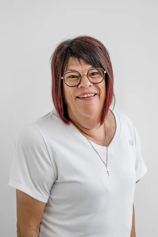 Eine lächelnde Frau mit kurzen dunklen Haaren und Brille, die ein weißes Hemd trägt, steht vor einem schlichten hellen Hintergrund.