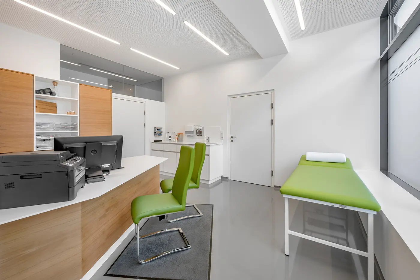 Moderne Arztpraxis mit grünen Stühlen, einem Schreibtisch mit Computern und einem Untersuchungstisch in der Nähe des Fensters.