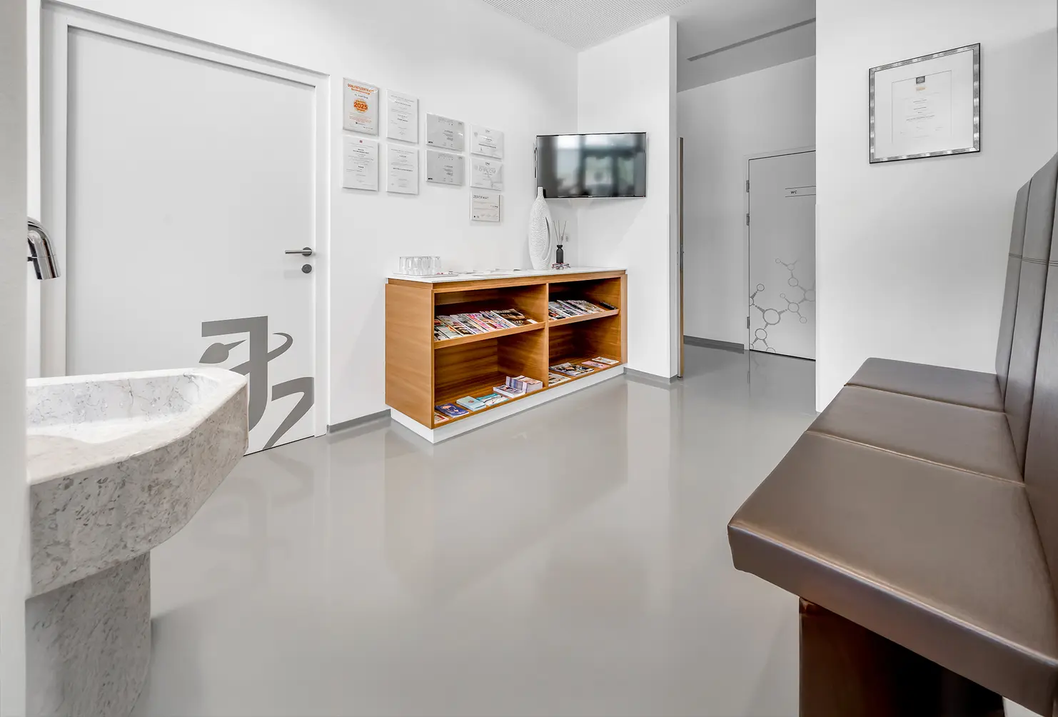 Modernes Wartezimmer mit einer Bank, einem Bücherregal mit Zeitschriften, einem an der Wand montierten Fernseher und einem Waschbecken auf der linken Seite.