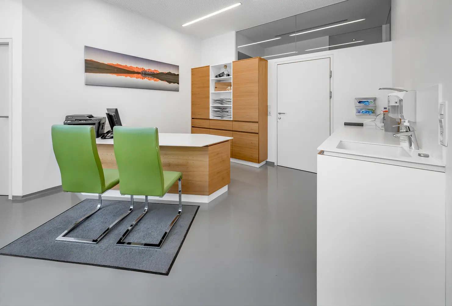 Ein modernes Büro mit zwei grünen Stühlen, einem Holzschreibtisch, Schränken und einem Wandfoto über dem Schreibtisch.