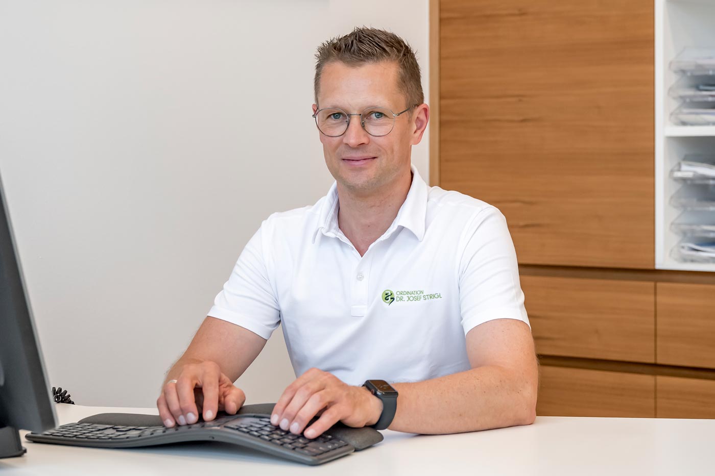 Ein Mann mit Brille und weißem Poloshirt arbeitet an seinem Schreibtisch und recherchiert lächelnd über Innere Medizin und Vorsorge.